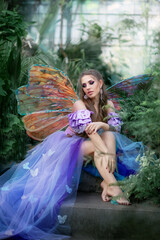 Fototapeta na wymiar A beautiful woman is a little cute fairy with butterfly wings in a green exotic forest. Fantasy shooting of fairy-tale characters. Forest nymph, pixie girl in purple dress