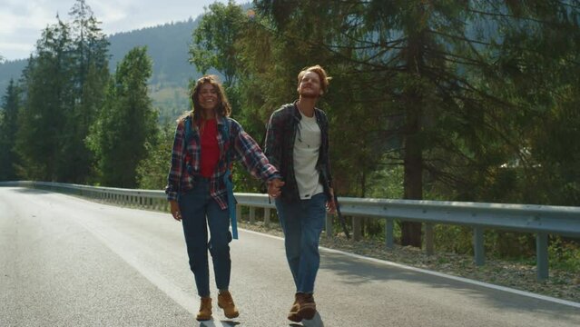 Happy walkers enjoy hitchhike in summer mountains. Hikers trekking on roadside.