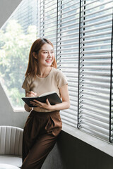 Smiling Asian woman holding a note in hand and standing near window Startup business Asian woman online SMEs Telemarketing