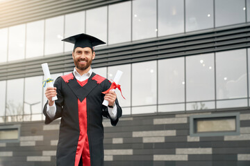 Front view of student with master degree standing, holding two diplomas, smiling. Handsome male in mortarboard and graduate gown graduating from high school. Concept of youth. - obrazy, fototapety, plakaty