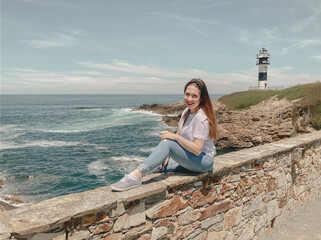 Young brown-haired woman, happy and smiling, sitting by a lighthouse facing the sea. Summer concept, holidays, tourism.