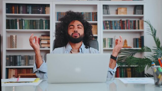 Young curly ethnic Arabian man freelancer takes lotus position from yoga after completing difficult task meditates to restore energy forces exhales and looks up sits at table with laptop in office