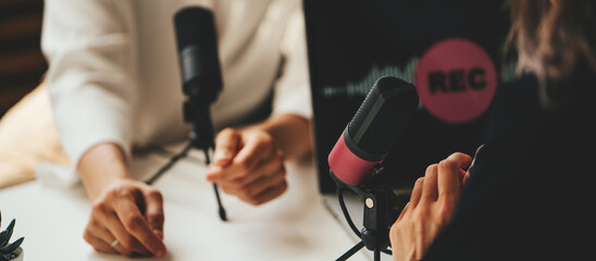 Two women streaming audio podcast using microphone and laptop at home broadcast studio, hands...