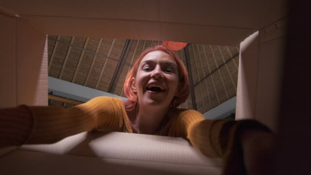 Young overjoyed attractive Caucasian woman millennial opens cardboard box and is delighted to see pleasant surprise stretches hands to get contents is happy to receive this gift be indoors