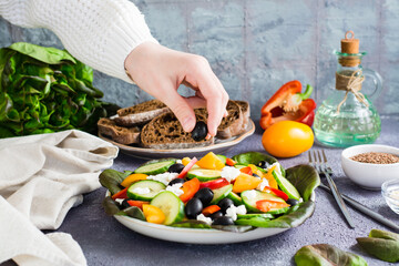 A woman's hand puts an olive in a freshly prepared Greek salad on a plate on the table.