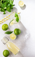 Flat-lay of mojito cocktail with lime, fresh mint and ice, bottle with mojito on white texture background with shadows. Summer refreshment citrus drink. Top view. Vertical orientation