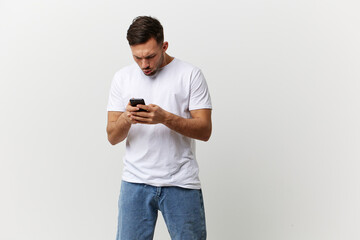 Angry confused irritated tanned handsome man in basic t-shirt type message with phone posing isolated on over white studio background. Copy space Banner Mockup. Distance online communication concept