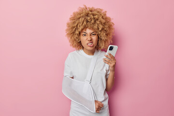 Photo of irritated curly haired woman frowns face uses mobile phone has arm dislocation wears sling...