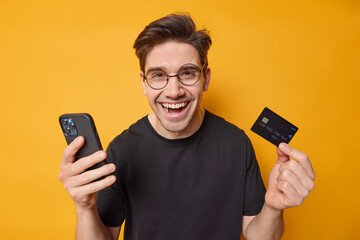 Happy dark haired man holds mobile phone and credit card presents how convenient to use mobile banking laughs joyfully wears round spectacles casual black t shirt isolated over yellow studio wall