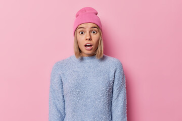 Waist up shot of shocked blonde European woman looks shocked and impressed stands speechless gasps from fear wears casual jumper hat isolated over pink studio background. Human reactions concept