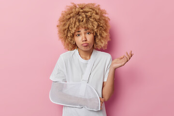 Waist up shot of unhappy woman with curly bushy hair puts on splint on broken arm shrugs shoulders doesnt know how to cure injury dressed in casual white t shirt isolated over pink background - obrazy, fototapety, plakaty