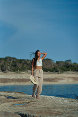 Young woman taking a barefoot walk on the beach enjoying the good summer weather, smiling with hat. Vertical photo.