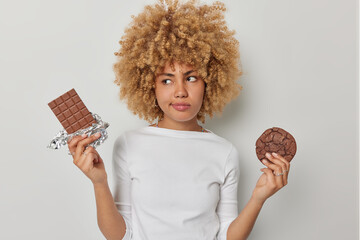 Thoughtful confused woman chooses between chocolate and cookie has sweet tooth dressed in casual...