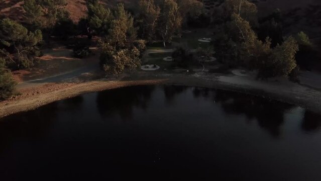 aerial footage of a vast still lake surrounded by lush green trees, grass and plants with majestic mountain ranges at Puddingstone Lake in San Dimas California USA