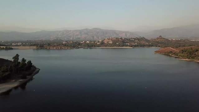aerial footage of a vast blue rippling lake at sunset surrounded by lush green trees, grass and plants with majestic mountain ranges and blue sky at Puddingstone Lake in San Dimas California USA