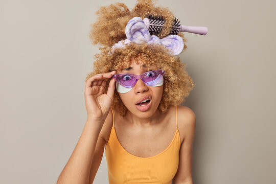 Anxious worried woman stares through purple shades notices something frightening undergoes beauty procedures applies white patches under eyes has comb stuck in curly hair isolated on grey wall
