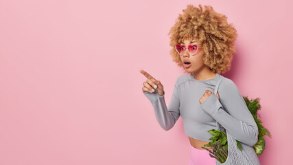 Horizontal shot of shocked curly haired young woman carries bag with fresh organic vegetables points index finger away on copy space isolated over pink background enjoys eating natural foods