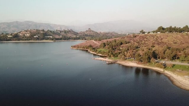 aerial footage of a vast blue rippling lake at sunset surrounded by lush green trees, grass and plants with majestic mountain ranges and blue sky at Puddingstone Lake in San Dimas California USA