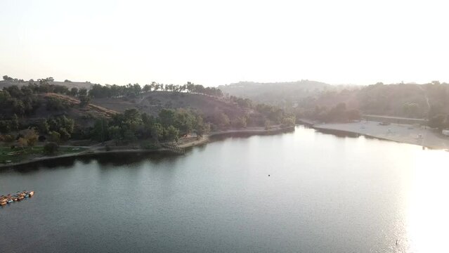 aerial footage of a vast blue rippling lake at sunset surrounded by lush green trees, grass and plants with majestic mountain ranges and blue sky at Puddingstone Lake in San Dimas California USA