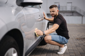Handsome man wipes a car with a rag in a showroom at a self-service car wash. Gray car. Polished...