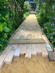 Close up of brick path construction of reclaimed clay yellow and red antique vintage house stones build straight garden pathway cutting through grass lawn and  plants and greenery under rose arch