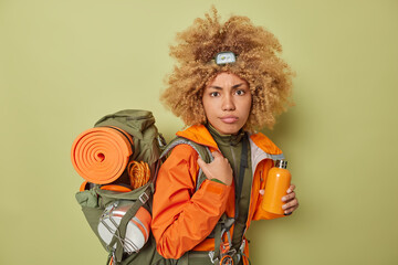 Serious female adventurer drinks water from bottle wears headlamp and orange jacket carries...