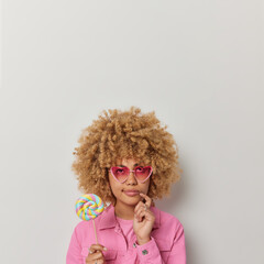 Pensive curly haired woman concentrated above being deep in thoughts holds multicolored lollipop thinks about something wears heart shaped sunglasses and pink jacket isolated over white wall