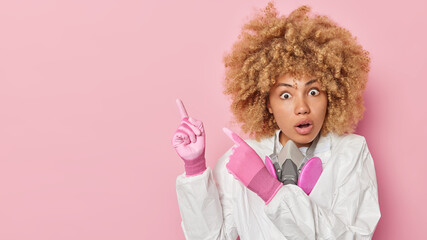 Horizontal shot of shocked woman in protective white suit and gloves points away on blank space shows danger tries to prevent you talks about chemical pollution empty space over pink background