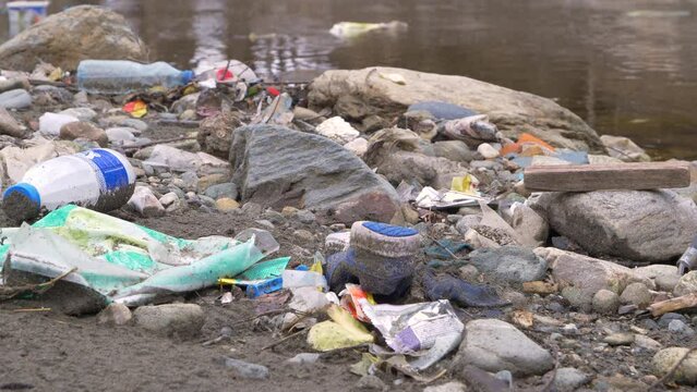 CLOSE UP: Flooded plastic rubbish and other trashes at the river bank as waste management issue. River debris and plastic garbage mix on a sandy river shore. The need to raise environmental awareness