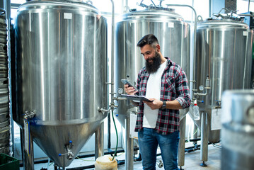 Fototapeta premium Man working in brewery and taking notes of the beer production and using smartphone