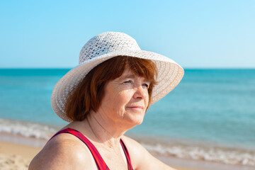 An elderly woman in a hat on the seashore on a sunny day, looks away and smiles, close-up. Summer pastime in retirement