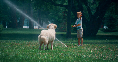 Little boy play water sprinkler on green field with playful golden retriever  - obrazy, fototapety, plakaty