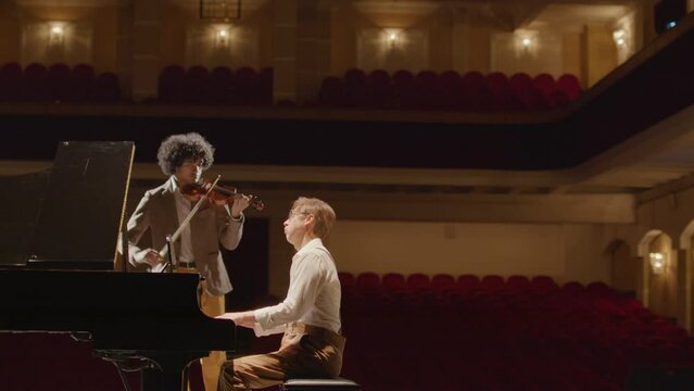 Wide shot of multiethnic duet playing violin and grand piano together on stage while preparing for classical music concert
