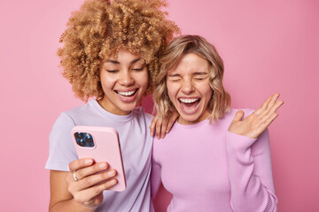 Overjoyed young women laugh happily watch video online hold mobile phone dressed in casual clothes...