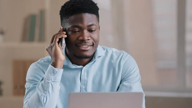 Smiling african man male businessman employee freelancer multitasking worker sit at home office talk on cellphone look at laptop screen provide remote communication business call use computer service