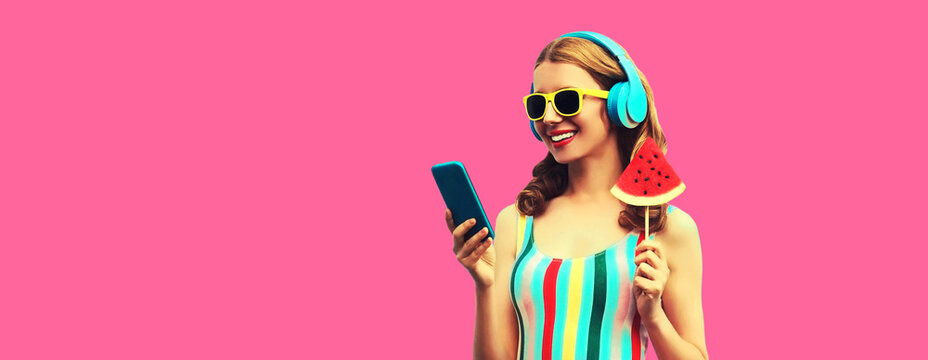 Summer portrait of happy smiling young woman model in headphones listening to music on smartphone with juicy lollipop or ice cream shaped slice of watermelon on pink background