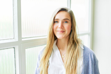 Portrait of young pretty 20 year old girl posing next to big window, looking straight at camera