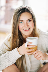Portrait of young pretty 20 year old girl relaxing at home with cup of tea or coffee, wearing white cozy cardigan, looking straight at camera