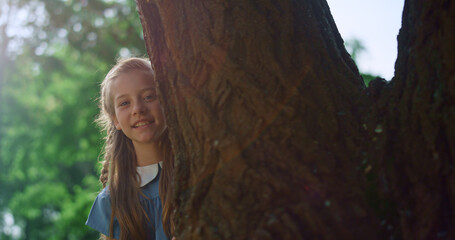 Cute blond girl looking out huge tree trunk. Child touching bark on sunny day.