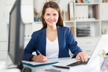 Portrait of confident smiling female office employee during daily work with laptop and documents