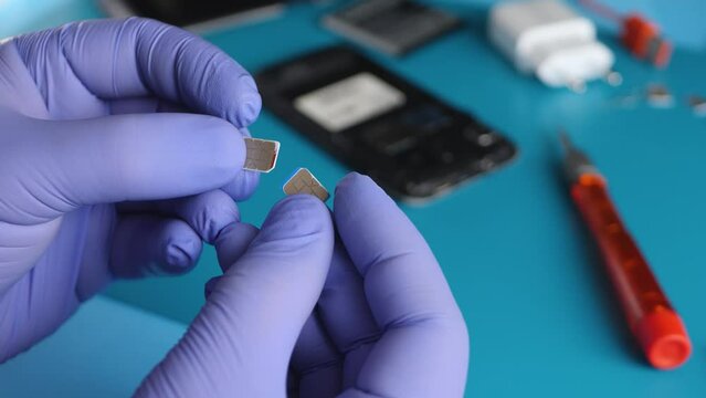 Unrecognizable hand holds nano SIM cards on disassembled mobile phone background. Communication equipment engineer in blue gloves in laboratory or factory