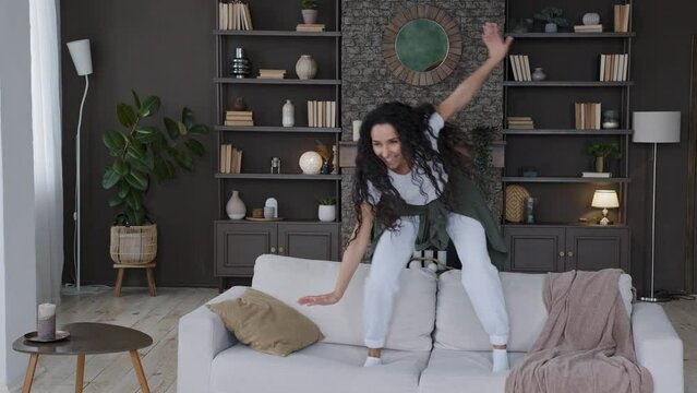 Young attractive fun cute joyful woman jumping on sofa in living room funny dancing actively comically moving to favorite music good mood enjoying weekend alone at home having fun makes comical moves