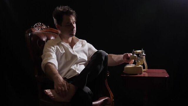 a man in a white shirt, trousers and barefoot picks up the phone in a retro phone and starts talking