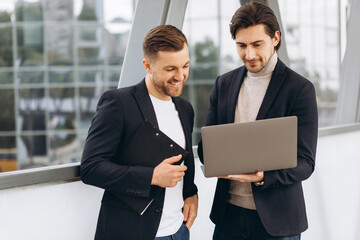 Two handsome young male businessmen in suits hold a laptop and a folder with documents and discuss a working project or have a video call against the background of urban office buildings.