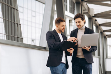 Two handsome young male businessmen in suits hold a laptop and a folder with documents and discuss a working project or have a video call against the background of urban office buildings.