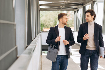 Two modern happy businessmen walking across a city bridge discussing something against the background of urban offices and buildings