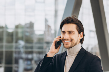 Portrait of handsome modern male businessman in suit talking by phone and smiling against the background of urban buildings and offices.