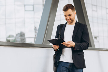 Portrait of handsome modern male businessman holding folder with documents on background of urban buildings and offices.