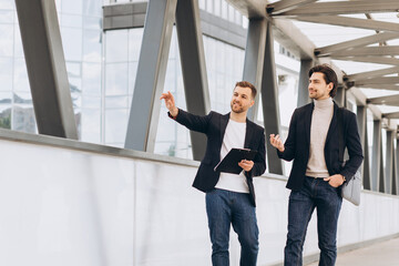 Two modern happy businessmen walking across a city bridge discussing something against the background of urban offices and buildings