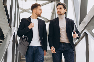 Two modern happy businessmen walking across a city bridge discussing something against the background of urban offices and buildings